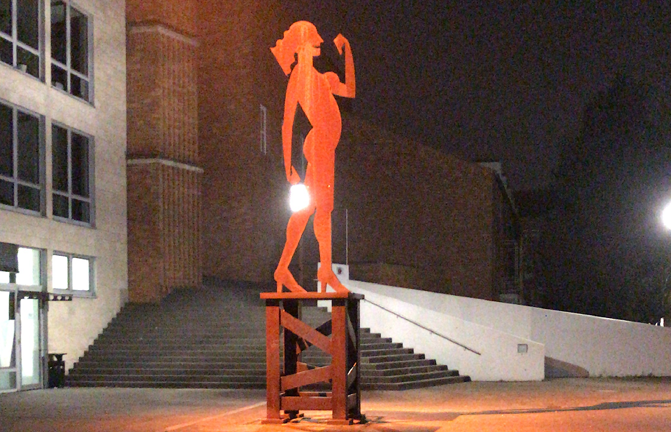 Zu sehen ist die Skulptur "Starke Frau" vor dem Stadthaus in Viersen, erleuchtet in orangefarbenem Licht
