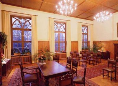 In einem historischen Raum mit teilweiser Holzverkleidung und großen Fenstern steht ein hölzerner Schreibtisch. Am Tisch stehen mehrere Stühle für die Zeremonie. Im Raum stehen noch weitere Stühle. Der Holzboden ist mit roten Teppichen ausgelegt und an der Decke hängen Kronleuchter.