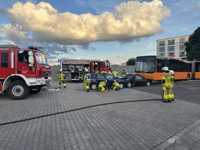 Das Bild zeigt mehrere Feuerwehrleute und Feuerwehrfahrzeuge sowie einen Linienbus, unter dem an der Front ein PKW eingeklemmt ist, in den offenbar ein weitere PKW von hinten hineingefahren ist. Die Feuerwehrleute arbeiten an den Fahrzeugen