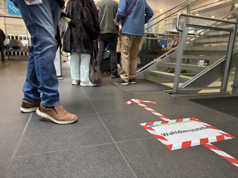 Das Bild aus dem Foyer des Stadthauses zeigt eine Bodenmarkierung "Wahldienststelle" sowie die Beine wartender Menschen